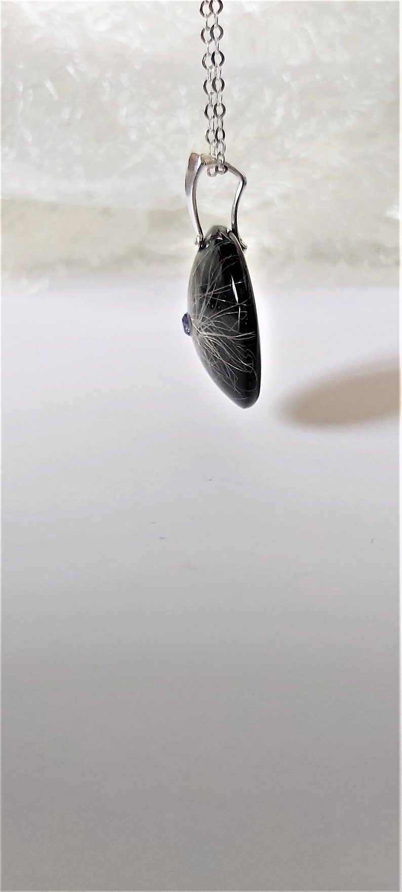 May include: A silver chain necklace with a black heart-shaped pendant containing a small blue gemstone and white hair.
