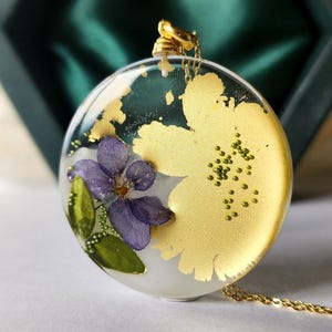Op de afbeelding: Een ronde hangerketting met een gouden ketting. De hanger is transparant met een gouden bloemenontwerp en een paarse bloem. Kleine groene kralen zijn ook zichtbaar in de hanger.