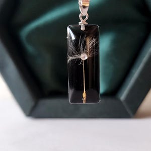 May include: A silver chain necklace with a rectangular black pendant. The pendant contains a dandelion seed head. The necklace is displayed in a dark green box.