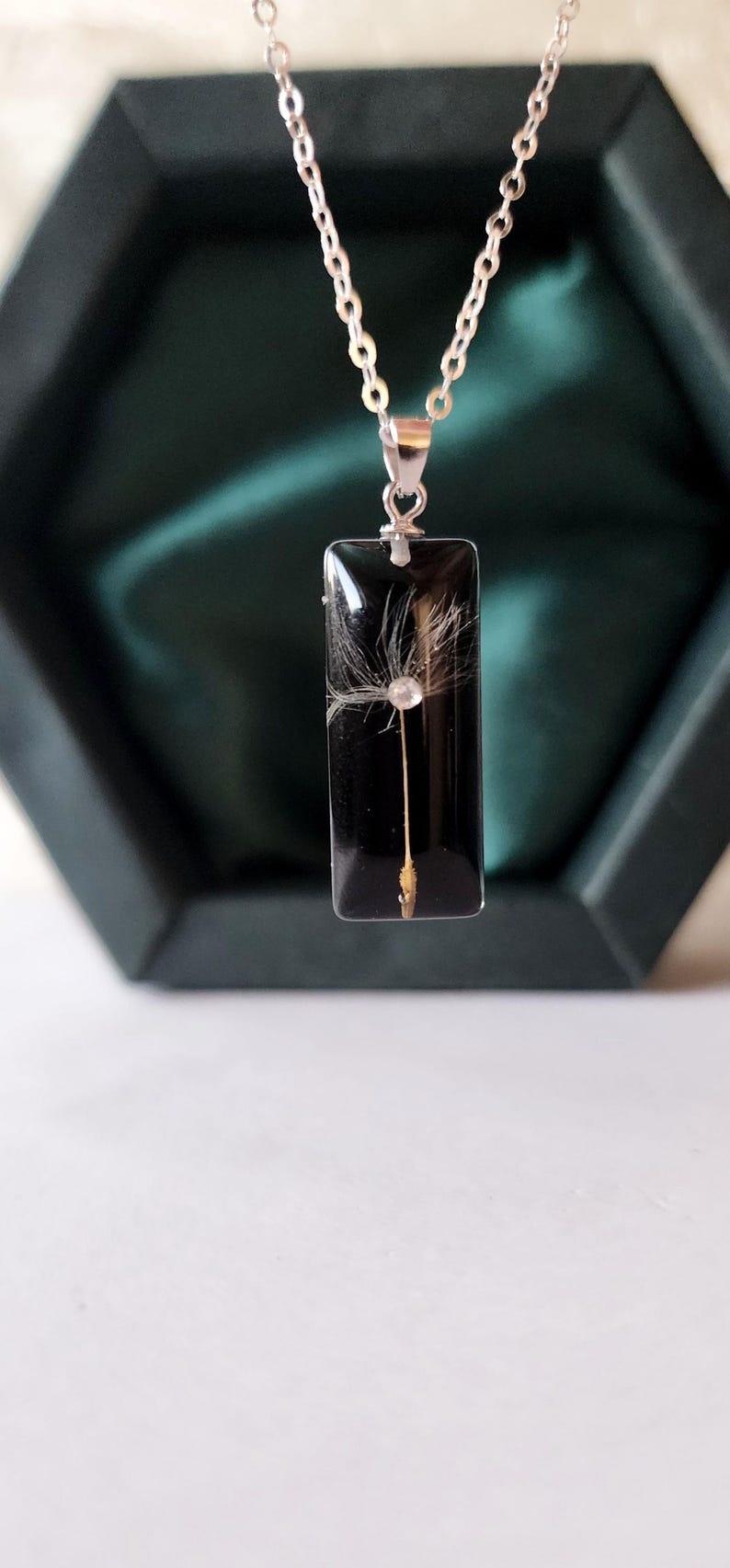 May include: A silver chain necklace with a rectangular black pendant. The pendant contains a dandelion seed head. The necklace is displayed in a dark green box.
