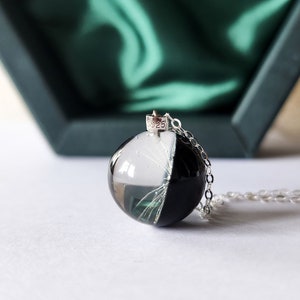 May include: A pendant necklace featuring a clear, round resin orb, split into black and white halves, with delicate white inclusions. The pendant hangs from a silver chain. The background includes a dark green box with a satin lining.
