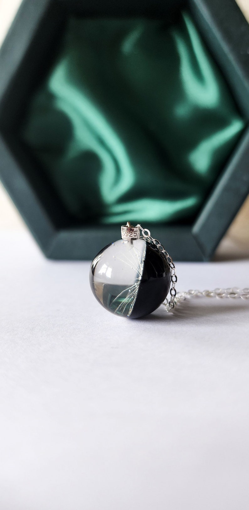May include: A pendant necklace featuring a clear, round resin orb, split into black and white halves, with delicate white inclusions. The pendant hangs from a silver chain. The background includes a dark green box with a satin lining.