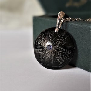 May include: A silver pendant with a black circular resin charm containing a small blue gemstone and strands of hair.