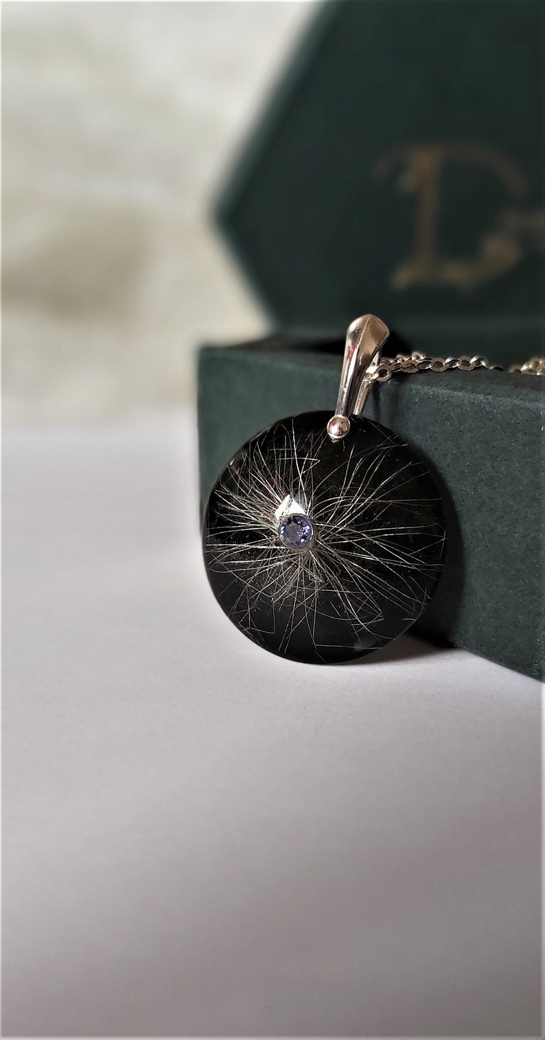 May include: A silver pendant with a black circular resin charm containing a small blue gemstone and strands of hair.