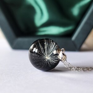 May include: A round, black pendant necklace with a silver chain. The pendant is a clear sphere containing a white dandelion seed head. The necklace is displayed on a white surface, with a green box in the background.