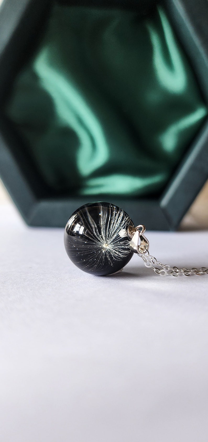 May include: A round, black pendant necklace with a silver chain. The pendant is a clear sphere containing a white dandelion seed head. The necklace is displayed on a white surface, with a green box in the background.