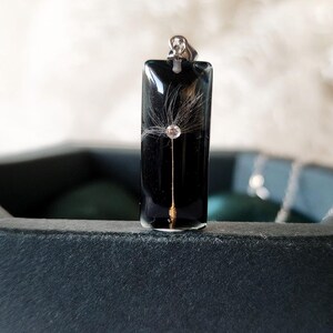 May include: A rectangular black pendant with a silver bail. Inside the pendant is a dandelion seed head with a small, clear crystal. The pendant is displayed on a dark surface.