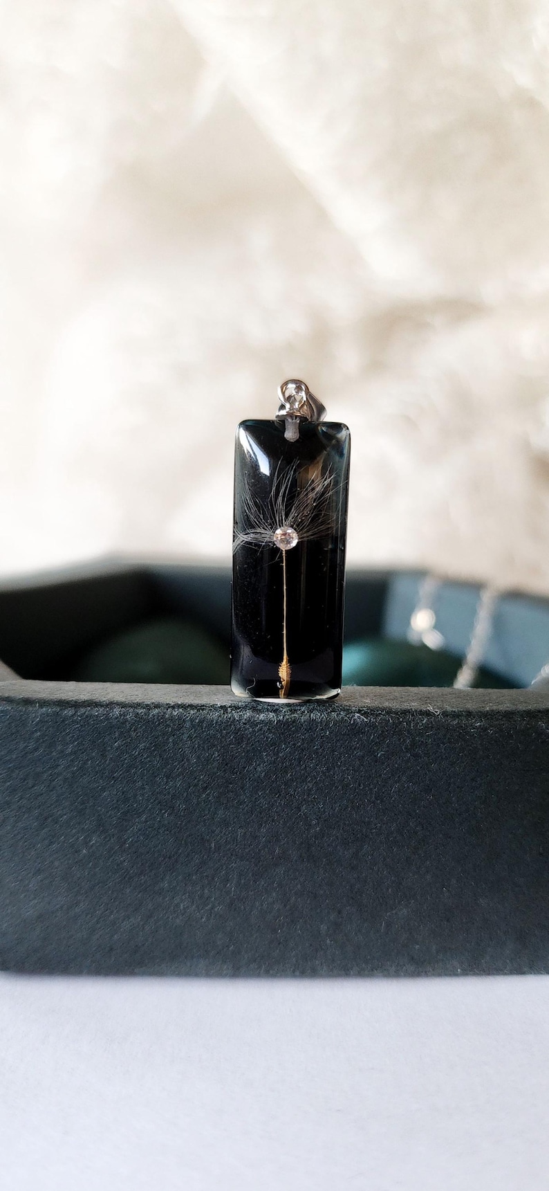 May include: A rectangular black pendant with a silver bail. Inside the pendant is a dandelion seed head with a small, clear crystal. The pendant is displayed on a dark surface.