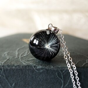 May include: A black resin pendant necklace with a dandelion seed design. The round pendant is suspended from a delicate silver chain. The necklace rests on a dark gray textured surface.