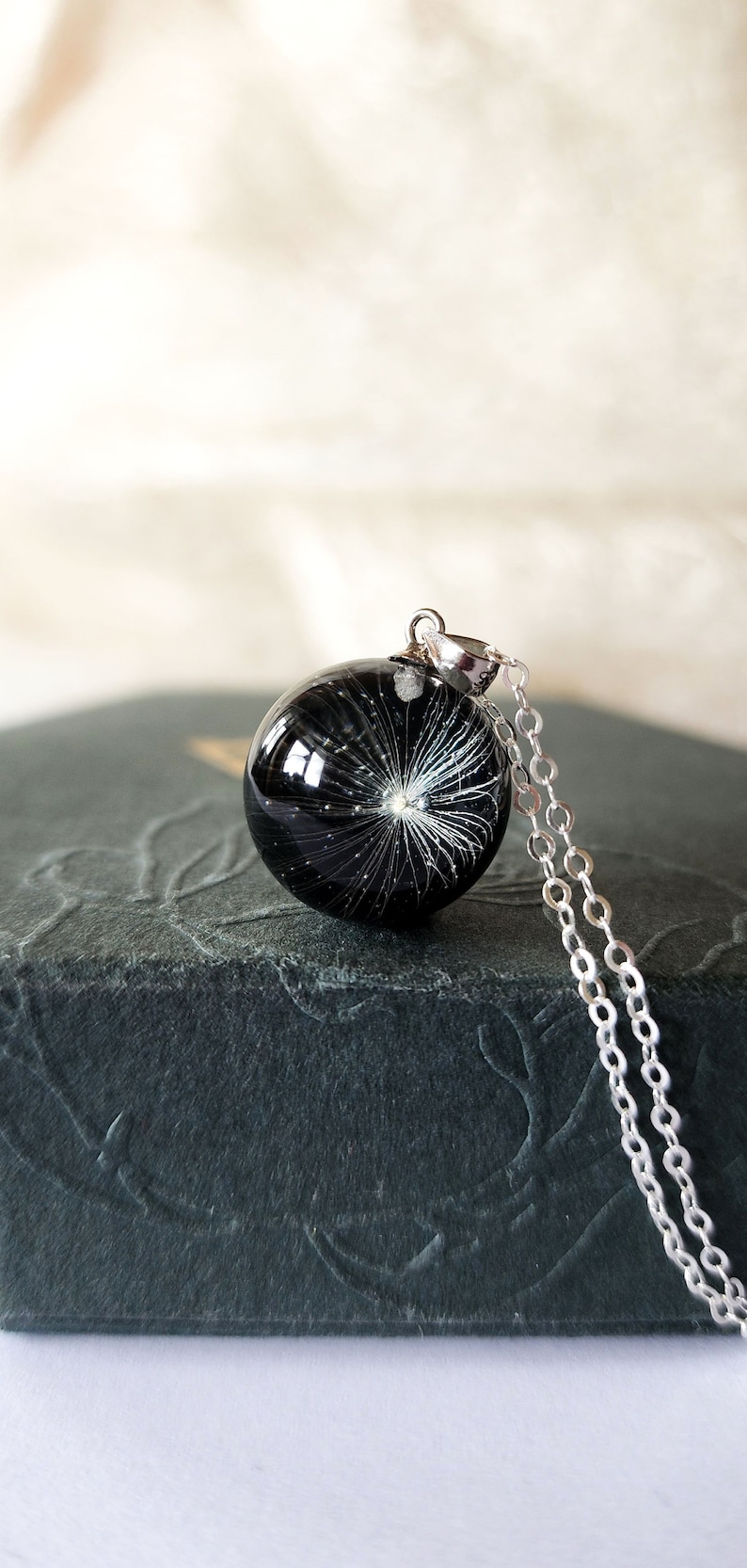 May include: A black resin pendant necklace with a dandelion seed design. The round pendant is suspended from a delicate silver chain. The necklace rests on a dark gray textured surface.