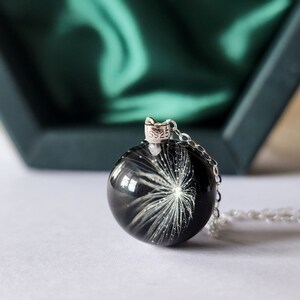 May include: A silver necklace with a round, black pendant. The pendant features a white, dandelion-like design. The necklace is displayed on a white surface with a green box in the background.