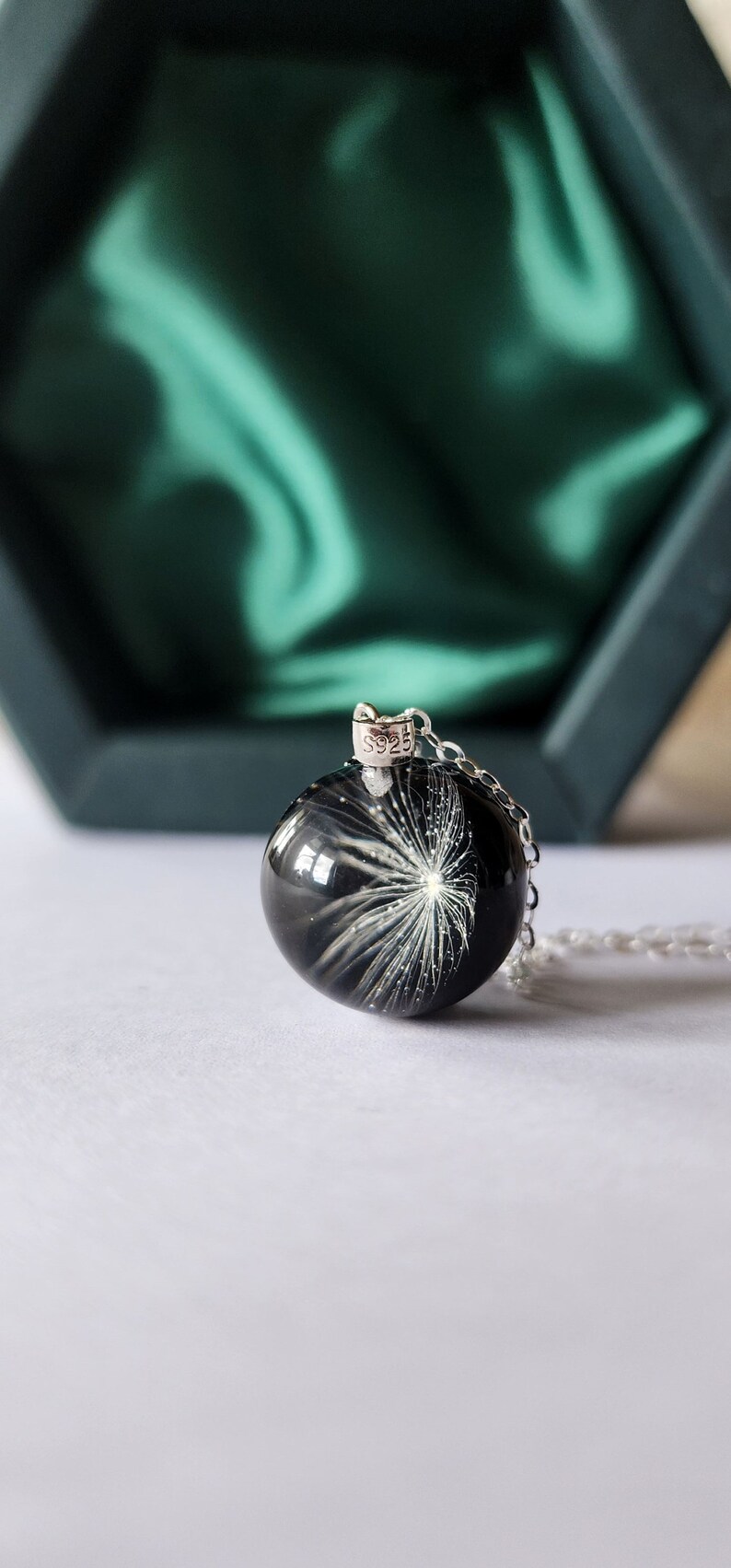 May include: A silver necklace with a round, black pendant. The pendant features a white, dandelion-like design. The necklace is displayed on a white surface with a green box in the background.