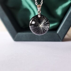 May include: A silver chain necklace with a round, black pendant. The pendant contains a white, feathery design, resembling a dandelion seed head. The necklace is displayed in a dark green jewelry box.