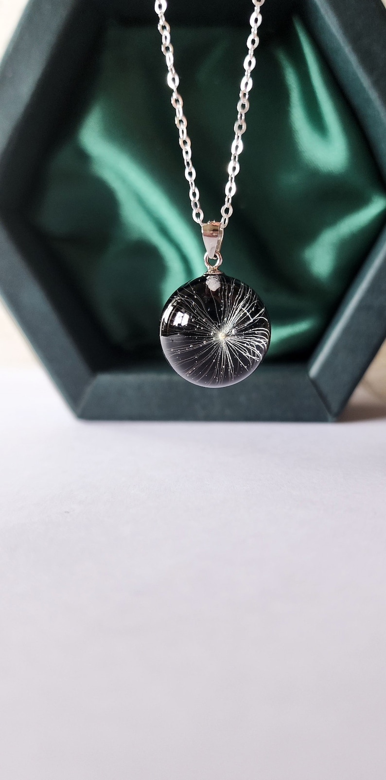 May include: A silver chain necklace with a round, black pendant. The pendant contains a white, feathery design, resembling a dandelion seed head. The necklace is displayed in a dark green jewelry box.