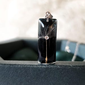 May include: A rectangular black pendant with a dandelion seed design. The pendant has a silver bail and a gold-colored stem. The dandelion seed is encased in clear resin. The pendant is displayed on a dark surface.