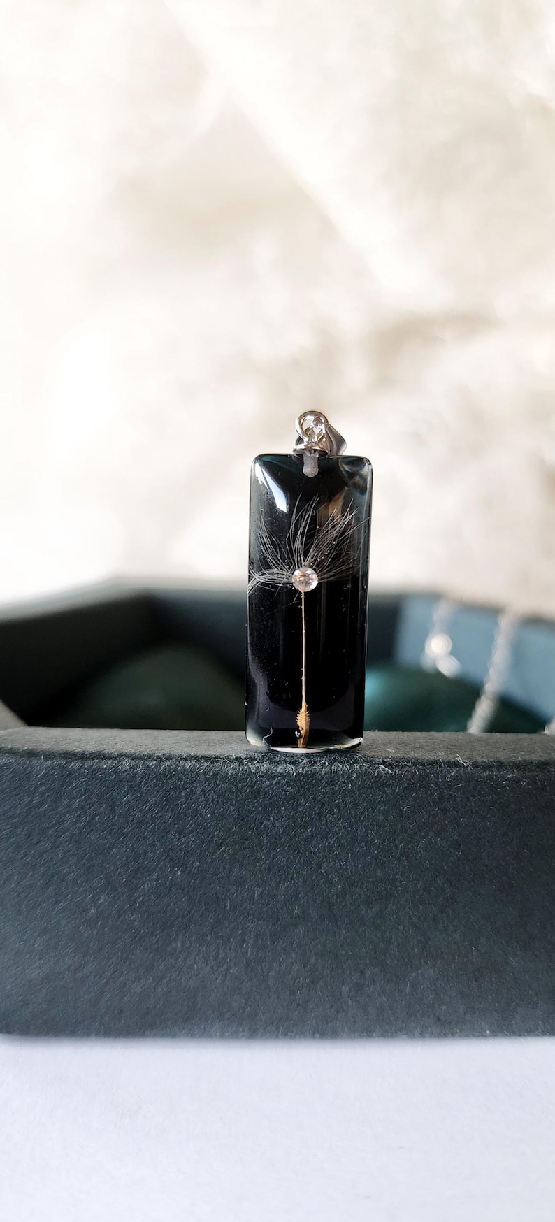 May include: A rectangular black pendant with a dandelion seed design. The pendant has a silver bail and a gold-colored stem. The dandelion seed is encased in clear resin. The pendant is displayed on a dark surface.