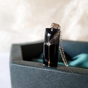 May include: A rectangular pendant necklace with a black background, featuring a delicate dandelion seed head encased in resin. The pendant hangs from a silver chain, presented in a dark gray jewelry box.
