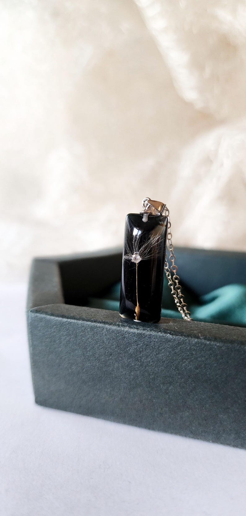 May include: A rectangular pendant necklace with a black background, featuring a delicate dandelion seed head encased in resin. The pendant hangs from a silver chain, presented in a dark gray jewelry box.