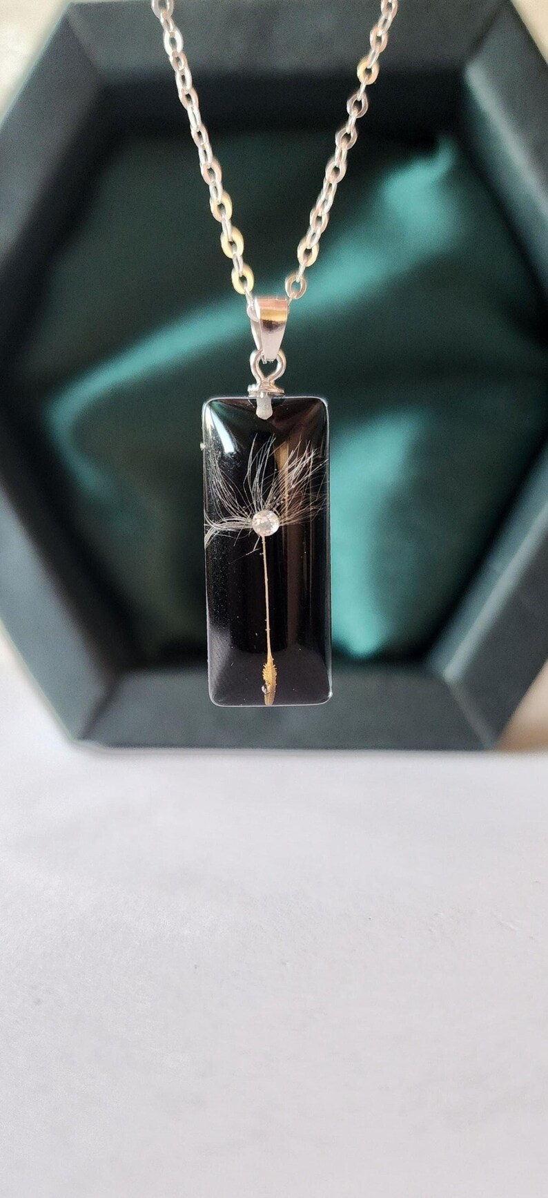 May include: A rectangular pendant necklace with a silver chain. The pendant is black with a dandelion seed head encased within. A small, clear crystal is set in the center of the seed head.