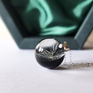 May include: A clear, spherical pendant necklace with a dandelion seed head encased in resin. The pendant hangs from a silver chain. The background includes a dark green box and a white surface.