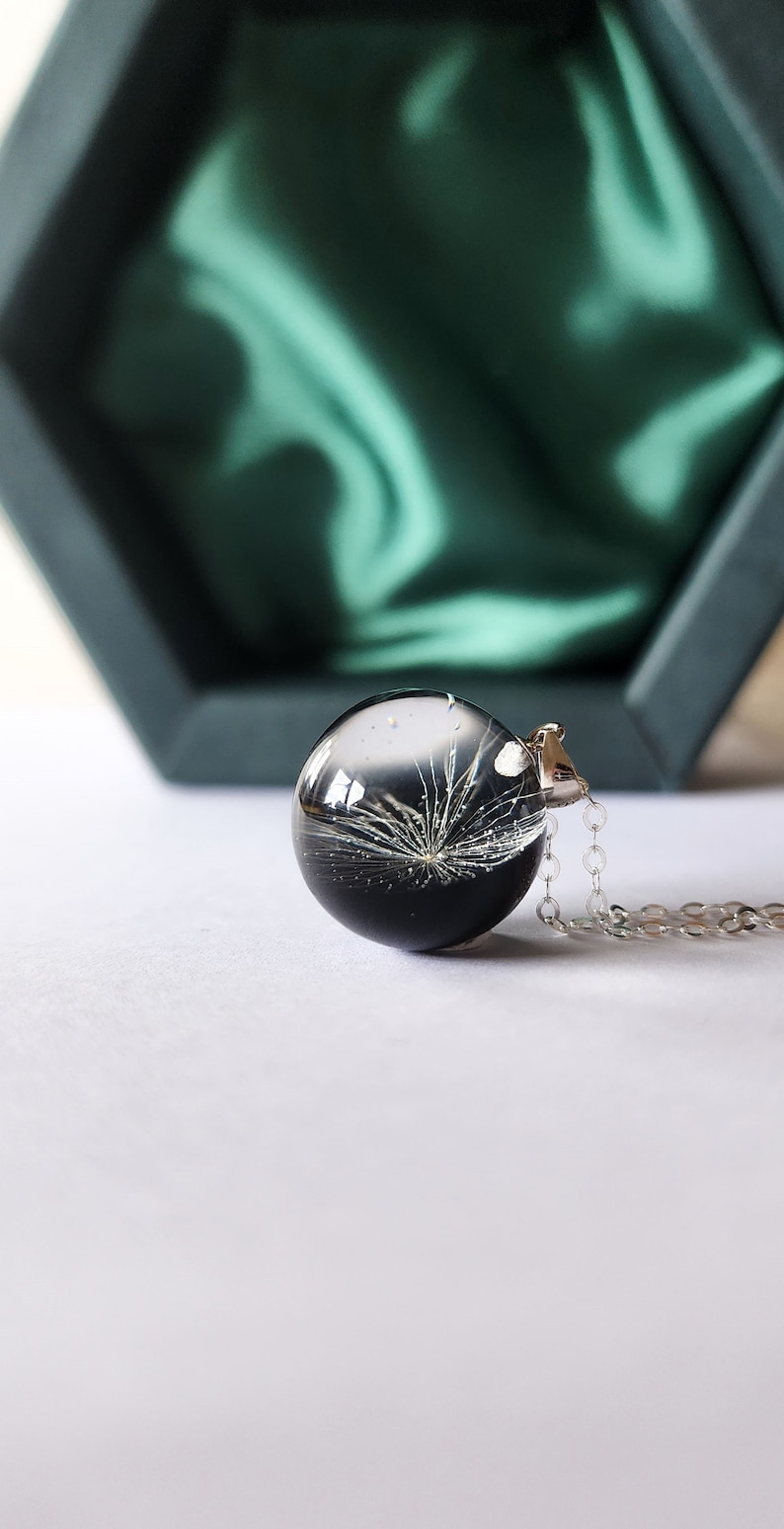 May include: A clear, spherical pendant necklace with a dandelion seed head encased in resin. The pendant hangs from a silver chain. The background includes a dark green box and a white surface.