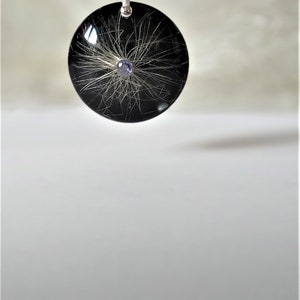 May include: A black round pendant with a silver chain. The pendant contains a small purple gemstone and white hair strands encased in resin.