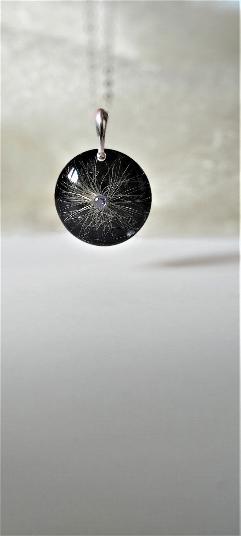 May include: A black round pendant with a silver chain. The pendant contains a small purple gemstone and white hair strands encased in resin.