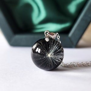 May include: A round, black pendant necklace with a silver chain. The pendant is a clear, glossy sphere containing a white dandelion seed head. The necklace is displayed on a white surface with a green box in the background.