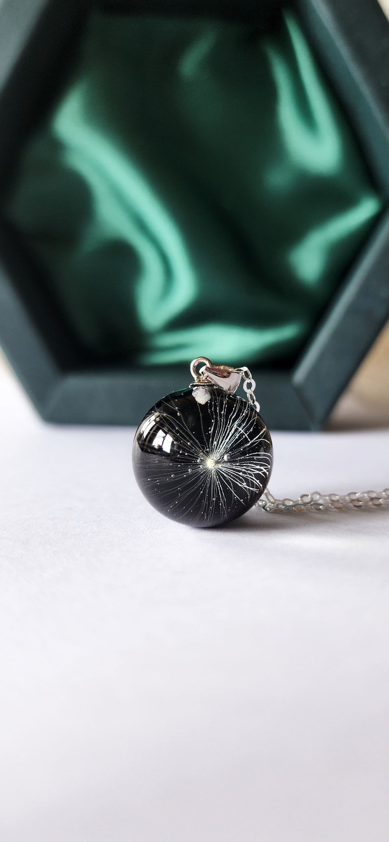 May include: A round, black pendant necklace with a silver chain. The pendant is a clear, glossy sphere containing a white dandelion seed head. The necklace is displayed on a white surface with a green box in the background.