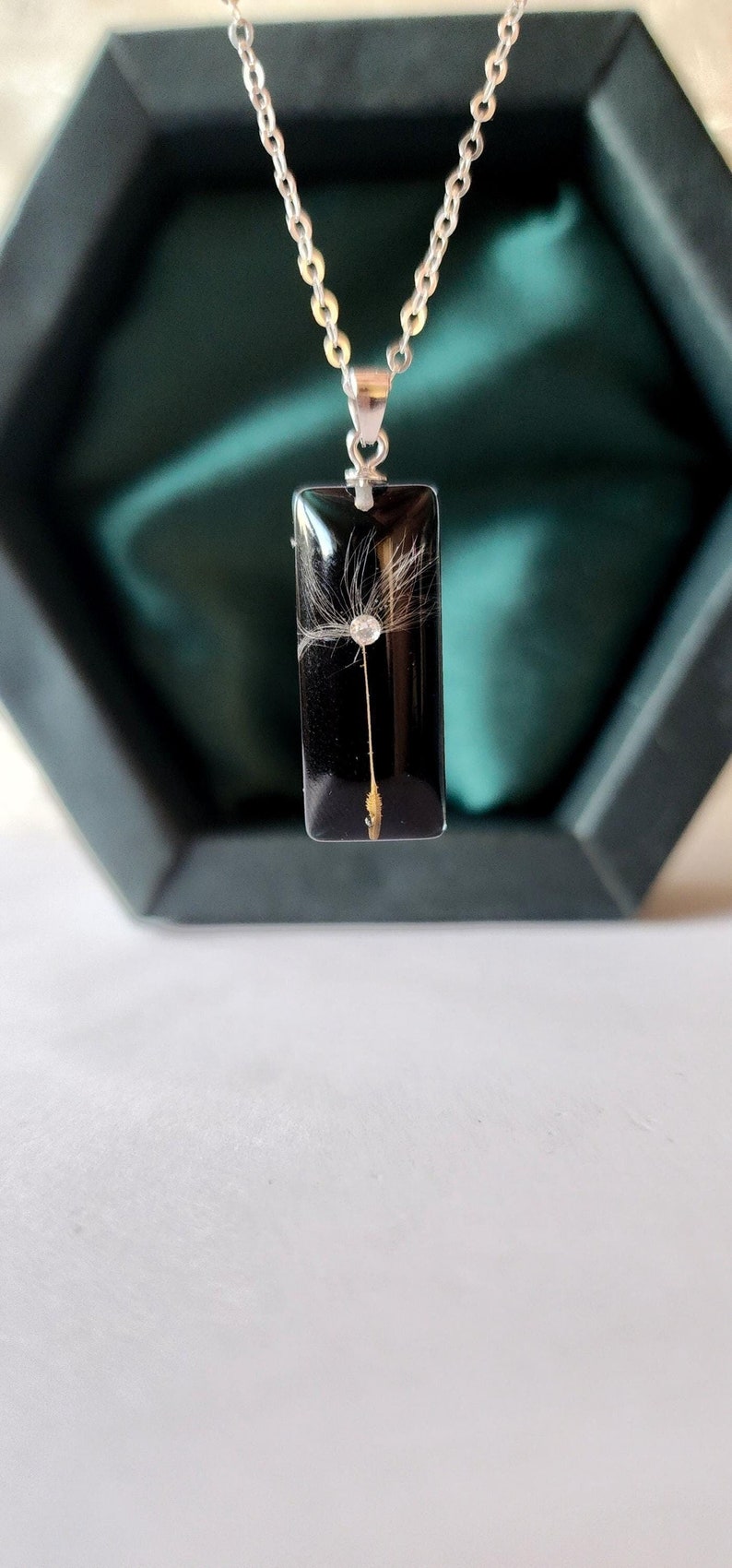May include: A silver chain necklace with a rectangular pendant. The pendant is black with a dandelion seed head encased within. The dandelion seed head is white and delicate. The necklace is displayed in a dark green box.