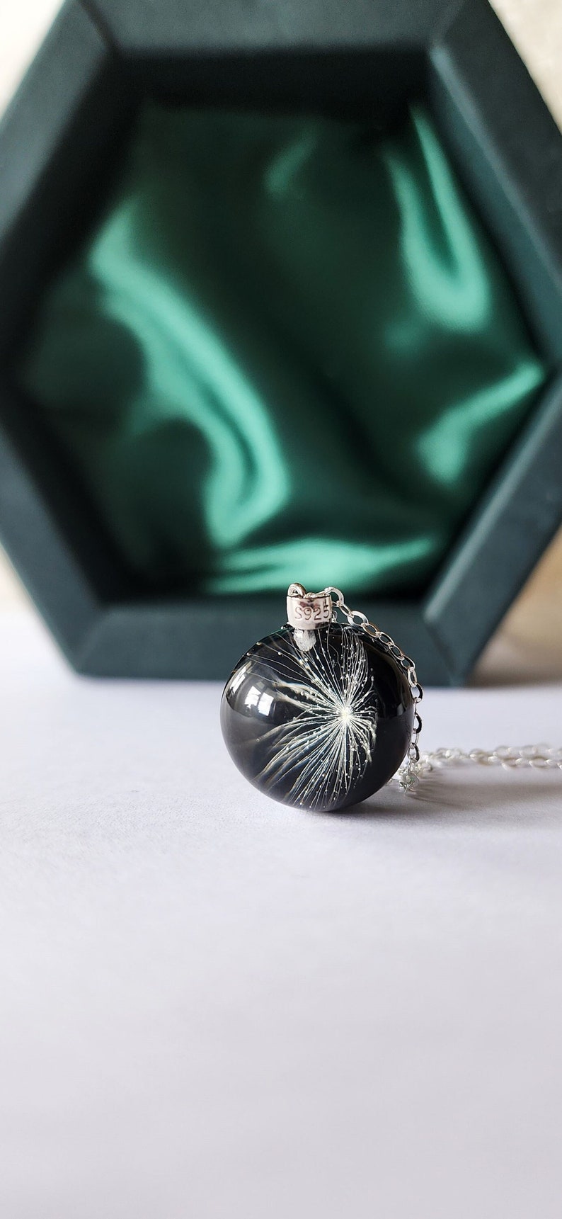 May include: A round, black resin pendant necklace with a silver chain. The pendant contains a white dandelion seed head. The necklace is displayed in front of a dark green box.
