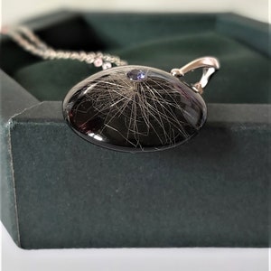 May include: A silver necklace with a black round pendant containing a small amount of hair and a purple gemstone.