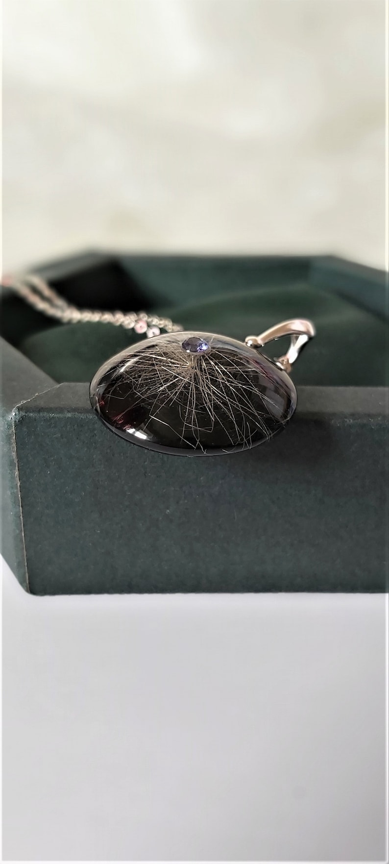 May include: A silver necklace with a black round pendant containing a small amount of hair and a purple gemstone.