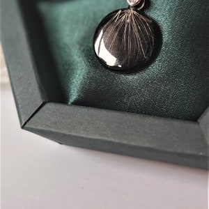Puede incluir: Un collar de plata con un colgante redondo que presenta un dise&ntilde;o de resina negra y un peque&ntilde;o acento de diamante. El collar se muestra en una caja de terciopelo verde.