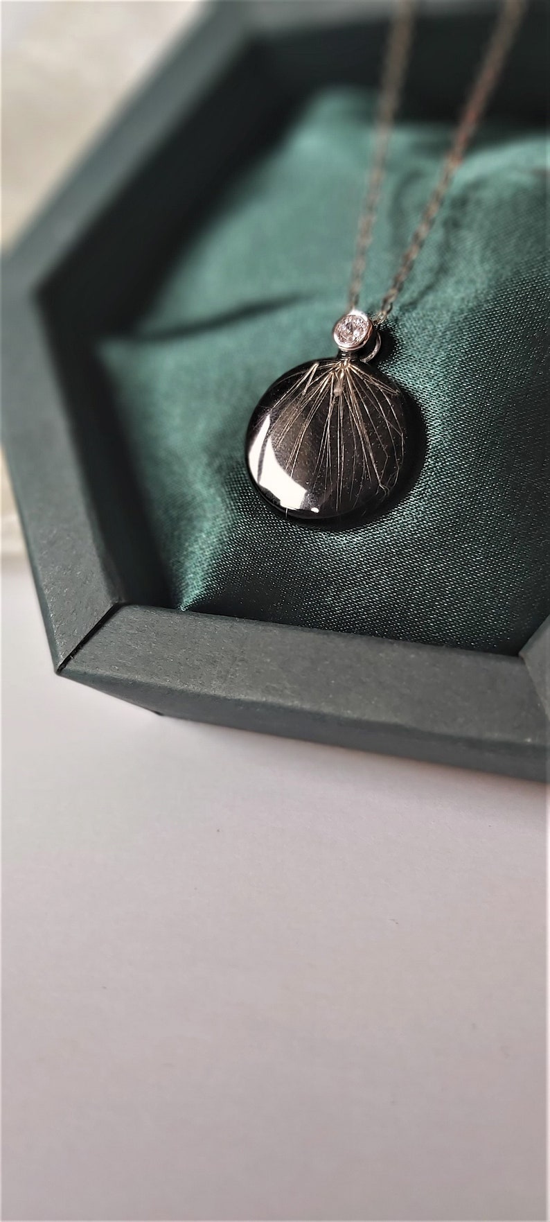 Puede incluir: Un collar de plata con un colgante redondo que presenta un dise&ntilde;o de resina negra y un peque&ntilde;o acento de diamante. El collar se muestra en una caja de terciopelo verde.