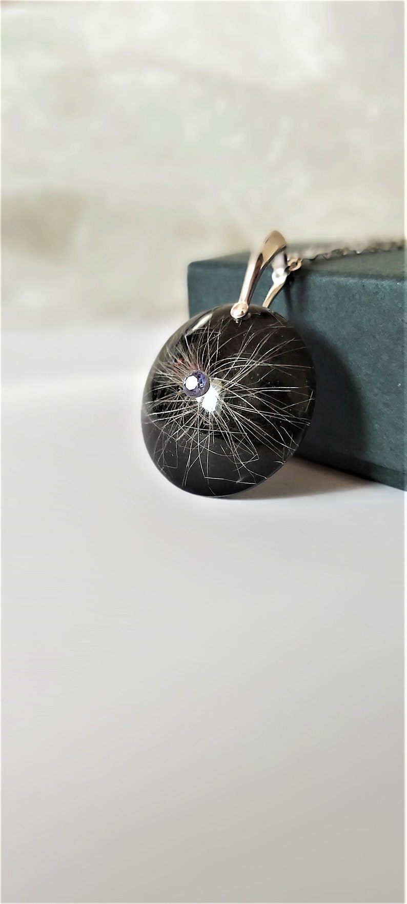 May include: A silver pendant necklace with a black resin round charm containing a small amount of hair and a small blue gemstone.