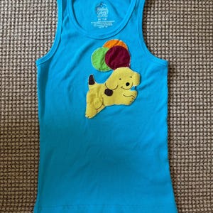 Pode incluir: Uma regata azul brilhante de tecido canelado com uma aplicação central de tecido de um cão amarelo com uma mancha castanha, segurando três balões coloridos em verde, laranja e bordeaux. A etiqueta interior mostra o texto Faded Glory e M 7-8.