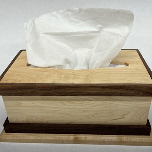 May include: A wooden tissue box cover with a maple wood top and walnut wood trim. The box has a rectangular shape and a slot for tissues.