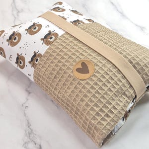Könnte beinhalten: Eine gefaltete Wickelauflage mit braunem Waffel-Außenmaterial und einem weißen Stoffinnenraum mit braunen Bärenillustrationen. Ein beiges Gummiband sichert die Auflage, und ein Lederaufnäher mit einem Herz ist angebracht.