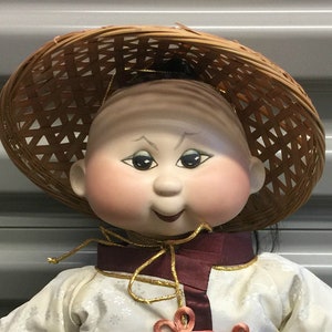 May include: A porcelain doll wearing a white and burgundy Chinese-style outfit with gold trim and a woven straw hat. The doll has a round face with rosy cheeks and black hair.