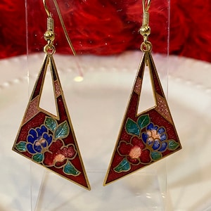 May include: A pair of gold tone dangle earrings with a geometric triangle shape. The earrings are decorated with red, pink, blue, and green enamel floral designs.
