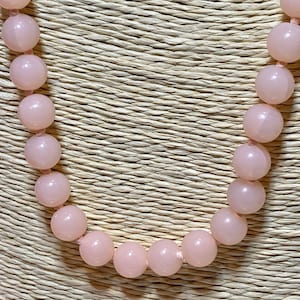 May include: A pink beaded necklace with round beads. The necklace is on a woven background.