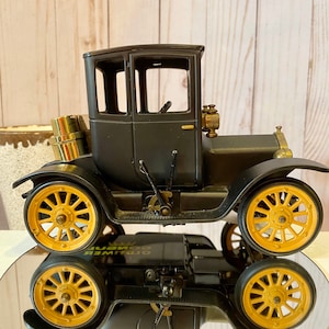 May include: Black vintage car model with gold wheels and a brass-colored gas tank. The car is on a reflective surface and the text "Oldtimer" is visible on the side.