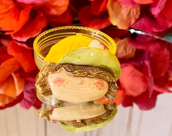 May include: A vintage plastic pin of a smiling girl wearing a yellow baseball cap. The pin is green, yellow, and pink.