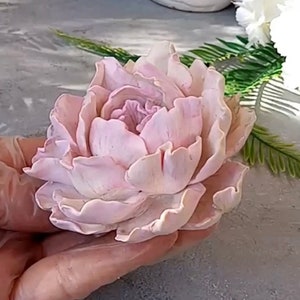 Pode incluir: Uma flor de pe&oacute;nia rosa p&aacute;lida delicada, feita &agrave; m&atilde;o com argila polim&eacute;rica. A flor tem um aspeto realista com detalhes intrincados e uma textura em camadas e com babados.