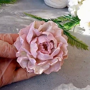 Pode incluir: Uma flor de pe&ocirc;nia rosa p&aacute;lida delicada, feita &agrave; m&atilde;o com argila polim&eacute;rica. A flor tem uma apar&ecirc;ncia realista com detalhes intrincados e uma textura macia e aveludada.