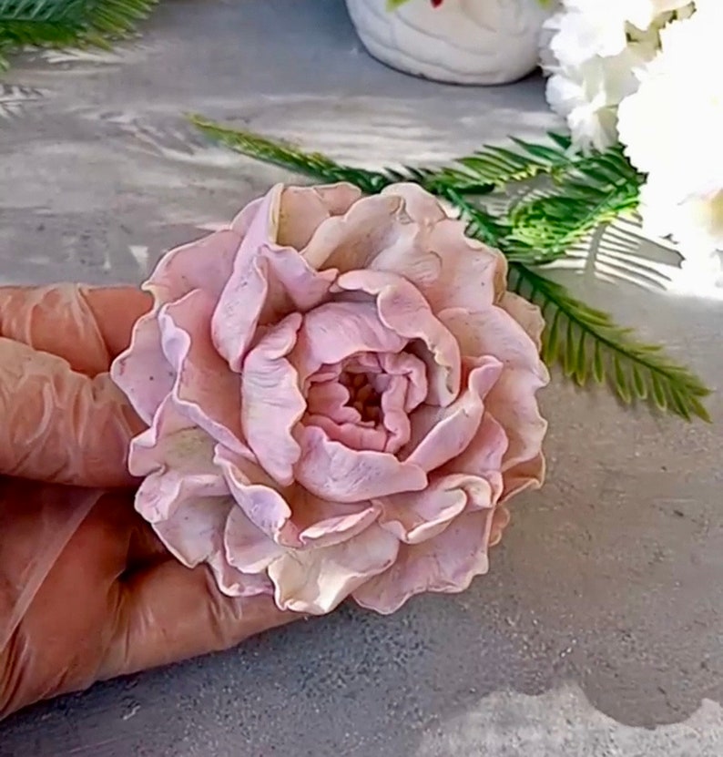 Pode incluir: Uma flor de pe&ocirc;nia rosa p&aacute;lida delicada, feita &agrave; m&atilde;o com argila polim&eacute;rica. A flor tem uma apar&ecirc;ncia realista com detalhes intrincados e uma textura macia e aveludada.
