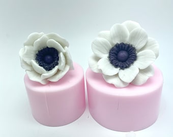 May include: Two white silicone molds with a white flower design. The flowers have a purple center and are sitting on top of the molds.