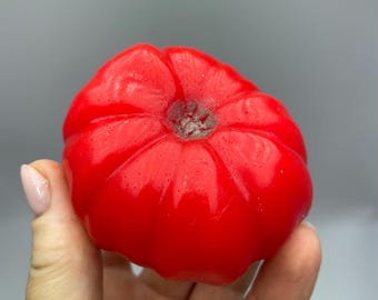 May include: A vibrant red, tomato-shaped candle. The candle has a glossy finish and detailed ridges, mimicking the natural form of a tomato. The candle is held in a hand against a gray background.
