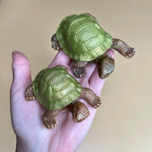 May include: Two small green turtle figurines with gold accents. The turtles are posed on their bellies with their legs outstretched.